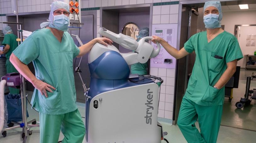 Prof. Rüdiger von Eisenhart-Rothe links und Dr. Florian Pohlig rechts mit dem Roboter-Assistenzsystem Mako in der Mitte im OP-Saal stehend  