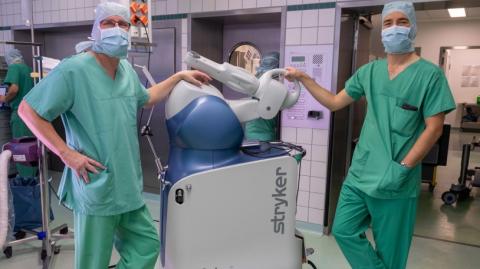 Prof. Rüdiger von Eisenhart-Rothe links und Dr. Florian Pohlig rechts mit dem Roboter-Assistenzsystem Mako in der Mitte im OP-Saal stehend 